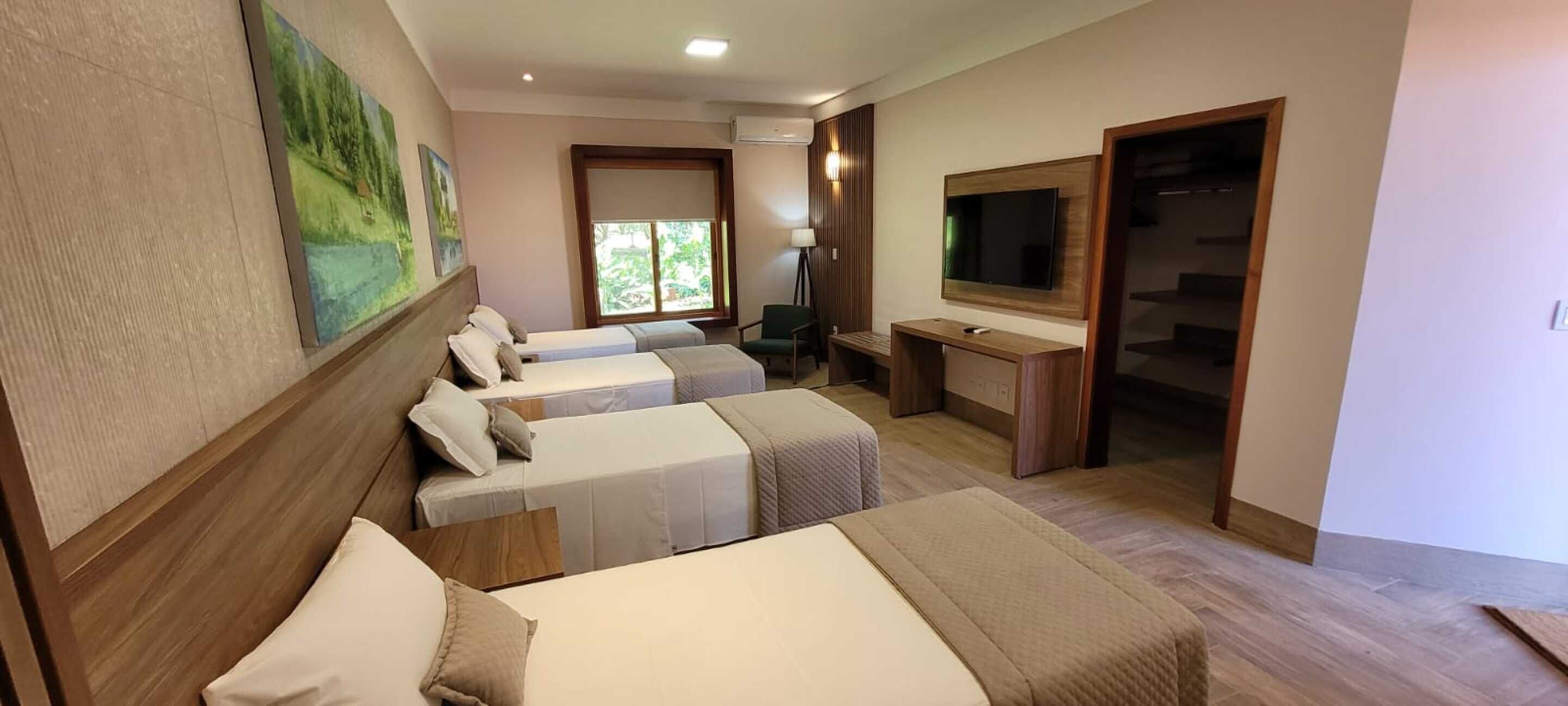 A hotel room with three single beds, a desk with a computer, a TV, and a large window.