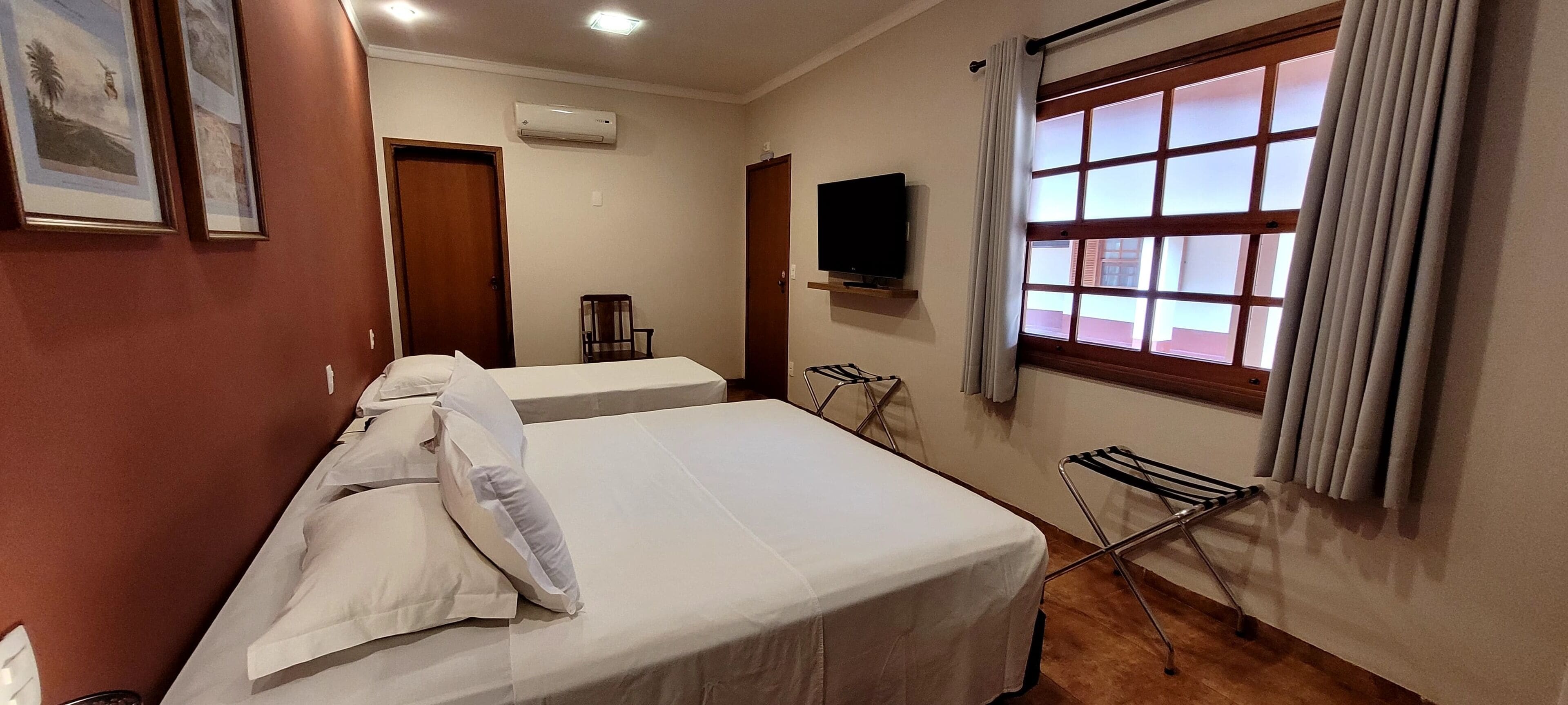 Quarto de hotel com duas camas, televisão, janela com cortinas e um escorredor de roupa.