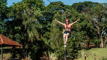 Uma pessoa praticando tirolesa por um parque verdejante, com árvores e uma estrutura de madeira ao fundo.