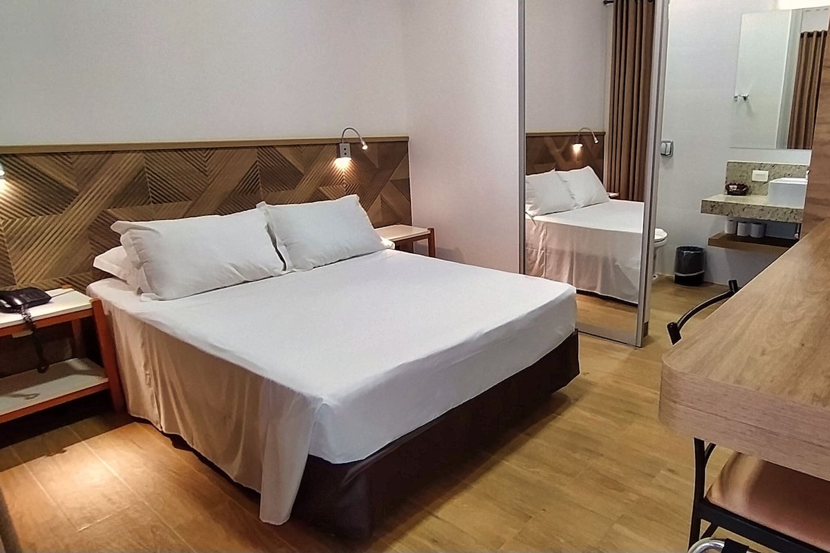 Habitación de hotel con cama, mesitas de noche, cabecero de madera, espejo, escritorio de madera y un pequeño cubo de basura.