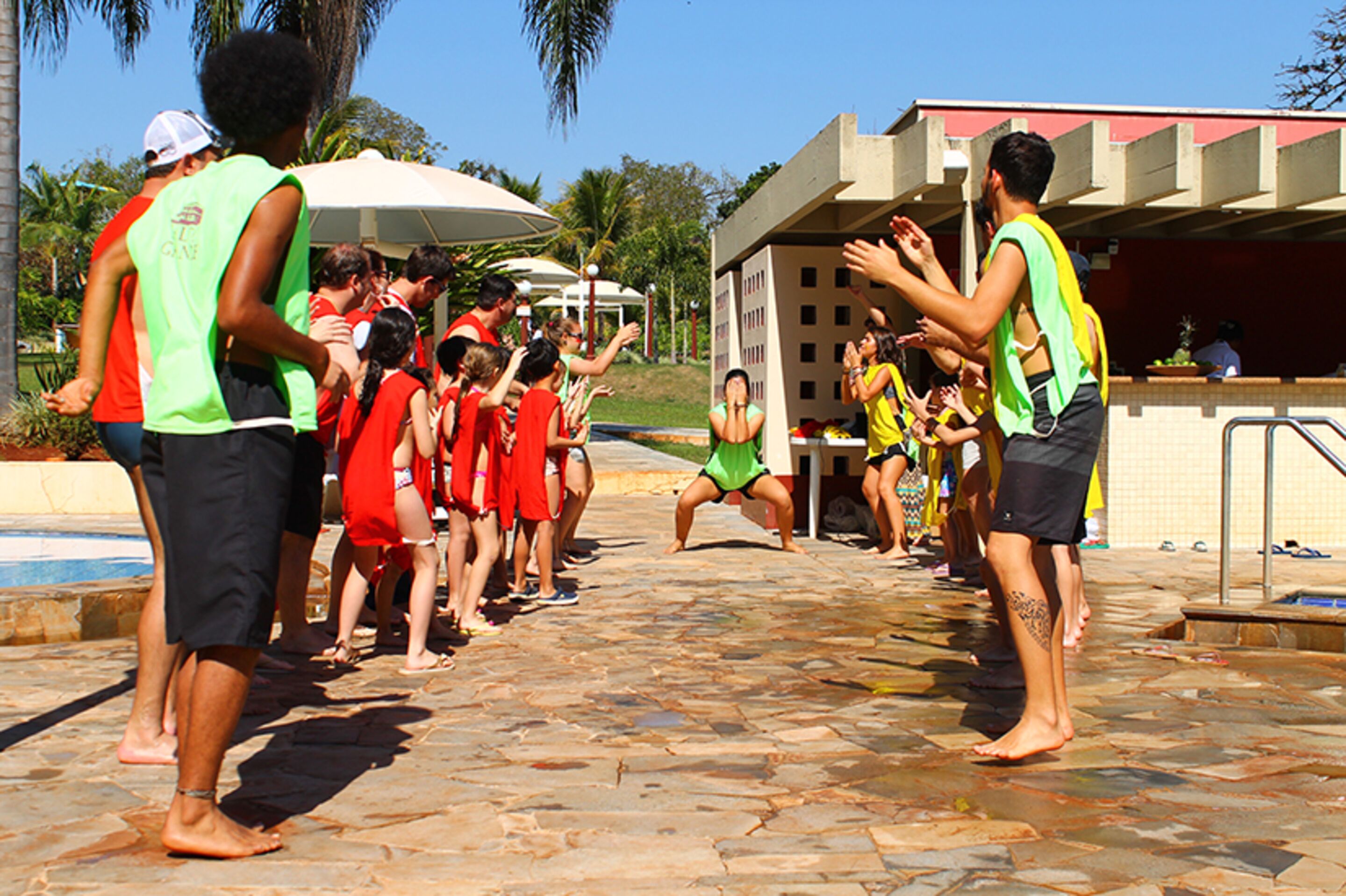 A group of people in a pool area with a stone floor, a covered seating area, and a building with a red roof.