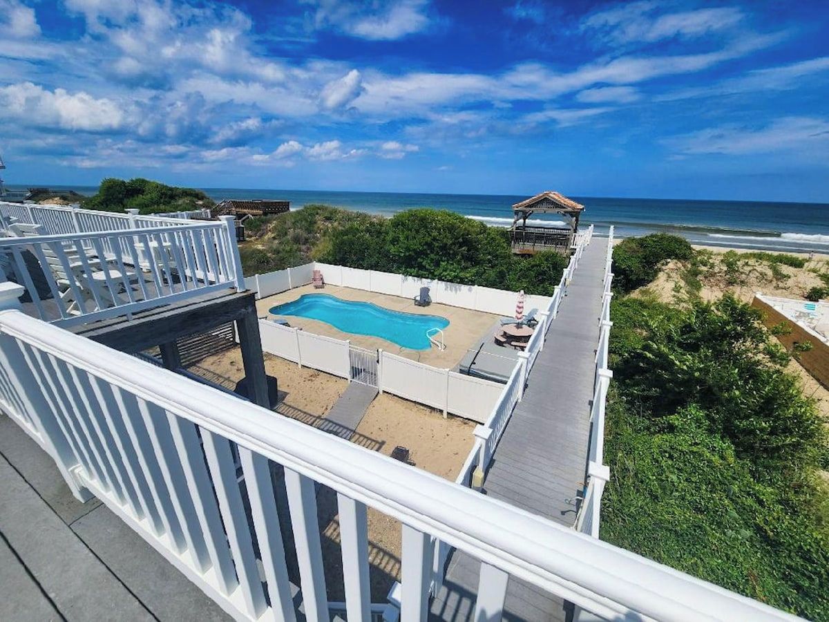 A rooftop pool with a view of the ocean, surrounded by greenery and a white fence.