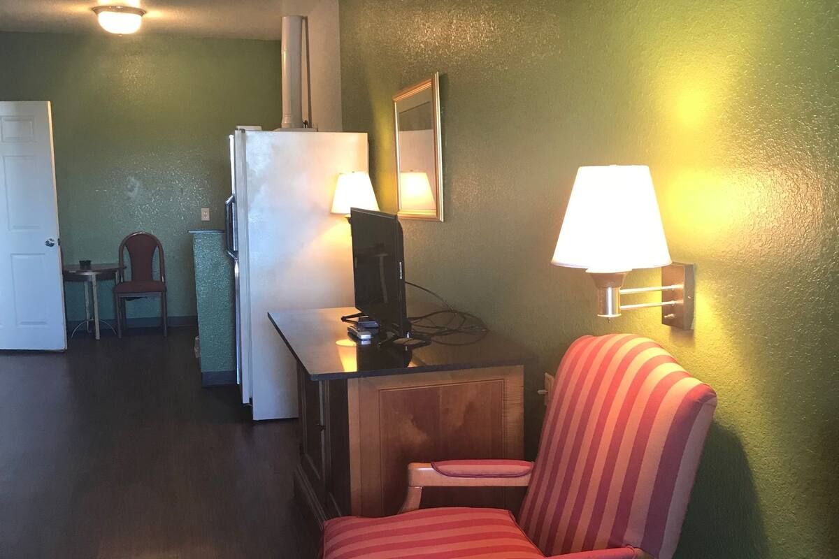 A room with a wooden desk, a striped armchair, a refrigerator, a lamp, and a mirror on the wall.
