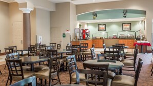 A hotel dining area with round tables and chairs, a kitchen in the background, and a reception desk.