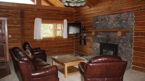 L’intérieur d’une cabane en rondins, avec une cheminée, des canapés en cuir, un téléviseur et un mur en pierre.