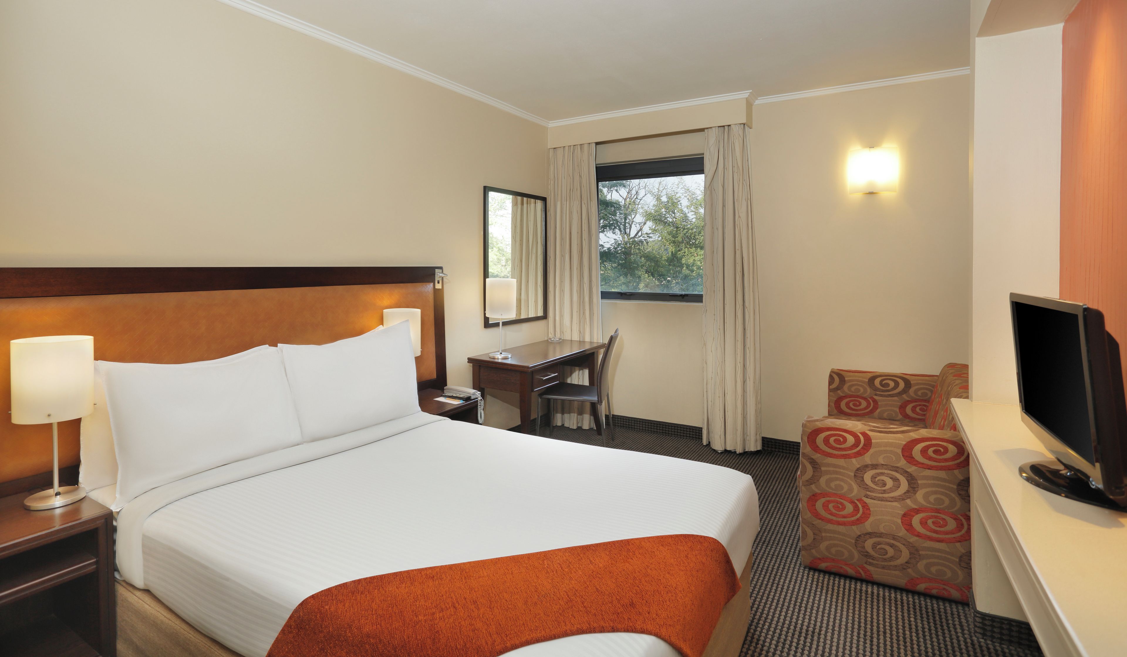 Habitación de hotel con cama, escritorio, silla, televisor y ventana con cortinas.