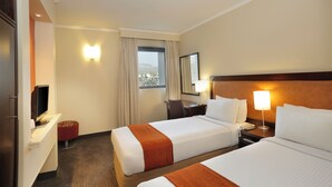 A hotel room with two beds, a desk, a TV, a window with curtains, and a view of the outside.