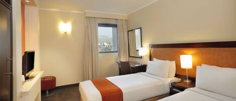 A hotel room with two beds, a desk, a TV, a window with curtains, and a view of the outside.