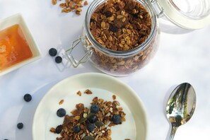 A glass jar filled with granola, a bowl of yogurt with granola and blueberries, and a spoon on a white surface. - Stayeasy Rustenburg (Rustenburg)