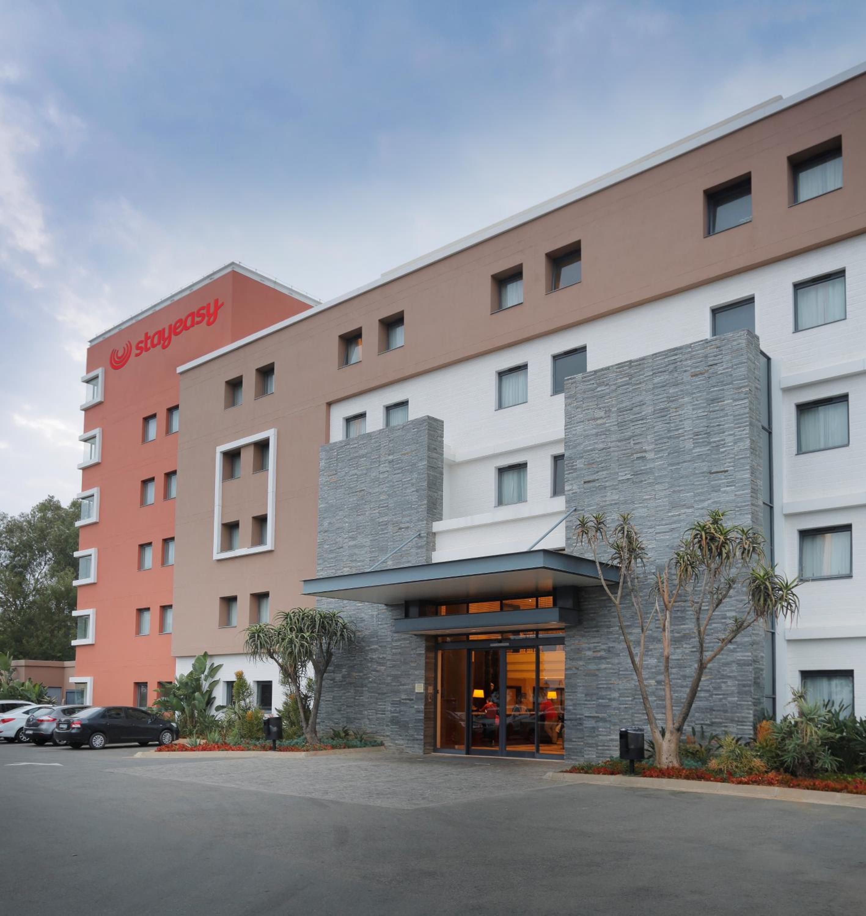 A multi-story hotel with a StayEasy sign, a parking lot, and a landscaped entrance area.