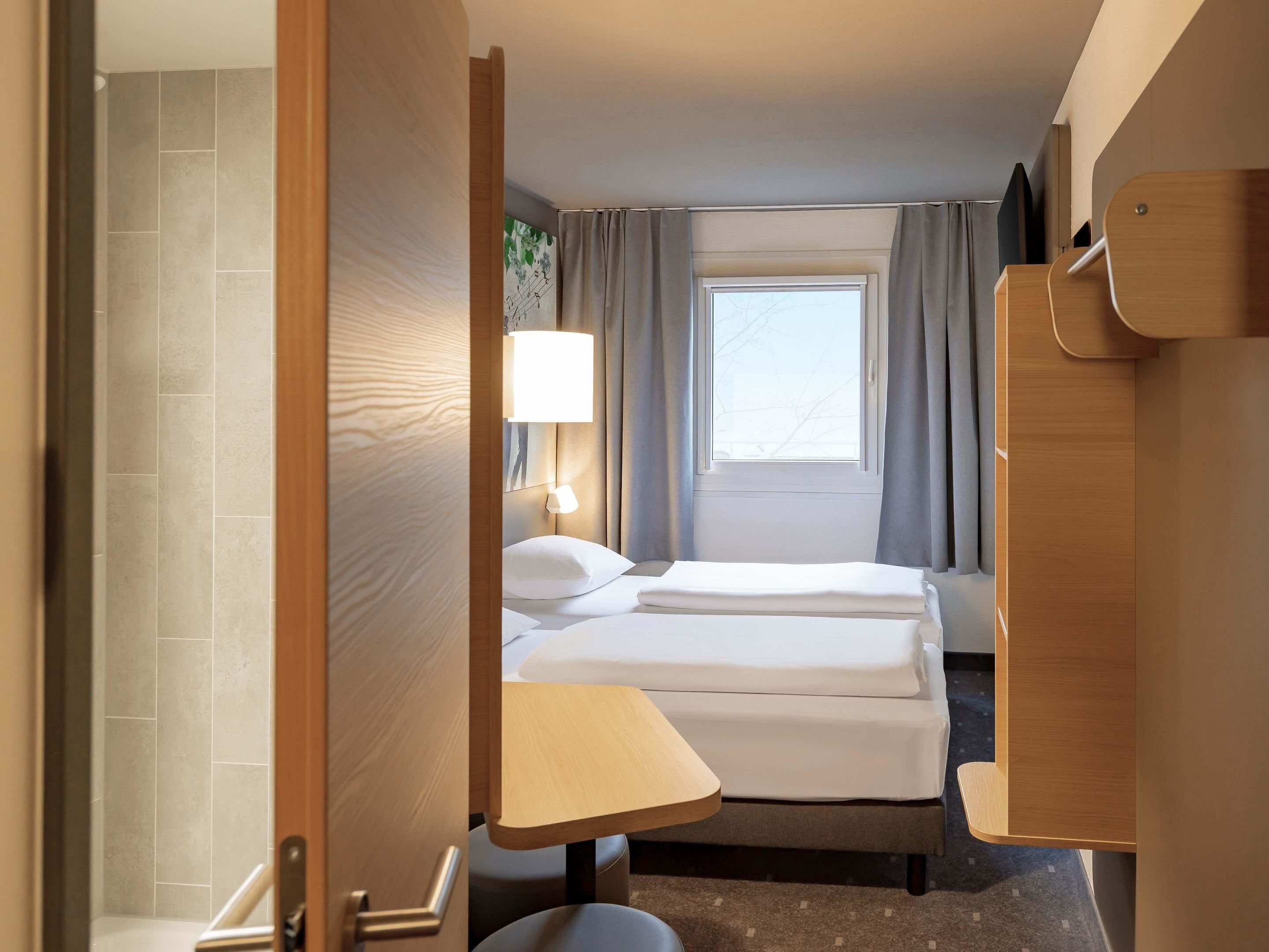 A hotel room with a bed, bedside table, lamp, window with curtains, and a door.