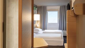 A hotel room with a bed, bedside table, lamp, window with curtains, and a door.