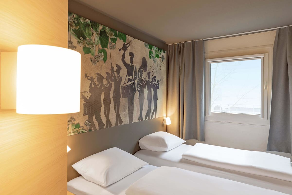 Habitación de hotel con dos camas, un mural en la pared, una ventana con cortinas y una lámpara de noche.