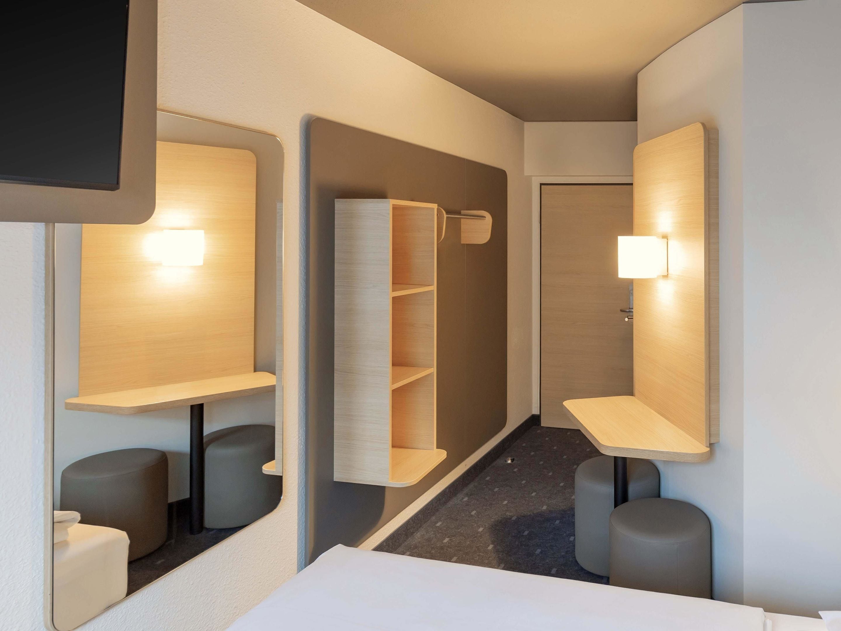 A modern hotel room with a minimalist design, featuring a large mirror, built-in shelves, and a small seating area.
