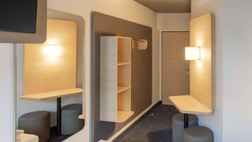 A modern hotel room with a minimalist design, featuring a large mirror, built-in shelves, and a small seating area.