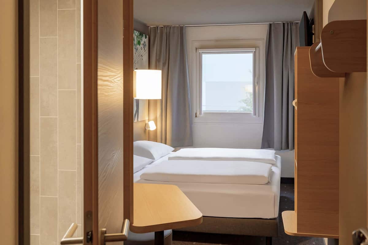 Una habitación de hotel con cama, mesita de noche, lámpara, ventana con cortinas y puerta.