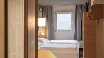 A hotel room with a bed, a bedside table, a lamp, a window with curtains, and a door.