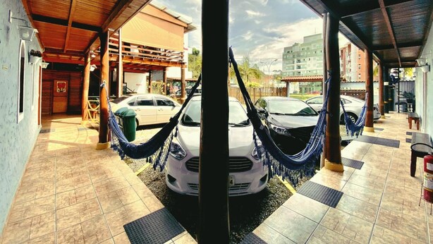 A covered parking area with a white car parked under a wooden structure, surrounded by tiled flooring and hammocks. - Pousada Beija Flor (Ubatuba)