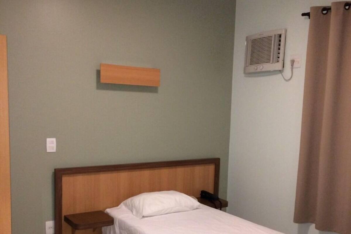 Un lit simple avec une tête de lit en bois, une table de chevet, un climatiseur mural et un rideau.