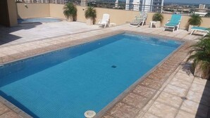 A rectangular swimming pool with a tiled edge, surrounded by lounge chairs and potted plants on a rooftop terrace.