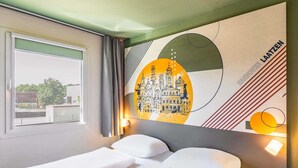 A hotel room with a bed, a window with curtains, a wall with a cityscape mural, and a nightstand with a lamp. - B&B HOTEL Hannover-Laatzen (Laatzen)