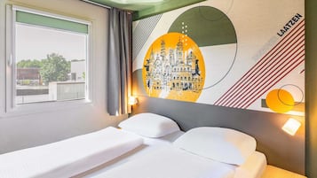 A hotel room with a bed, a window with curtains, a wall with a cityscape mural, and a nightstand with a lamp.