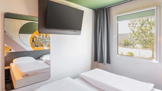 A hotel room with a bed, a TV mounted on the wall, a window with curtains, and a mirror reflecting the room. - B&B HOTEL Hannover-Laatzen (Laatzen)