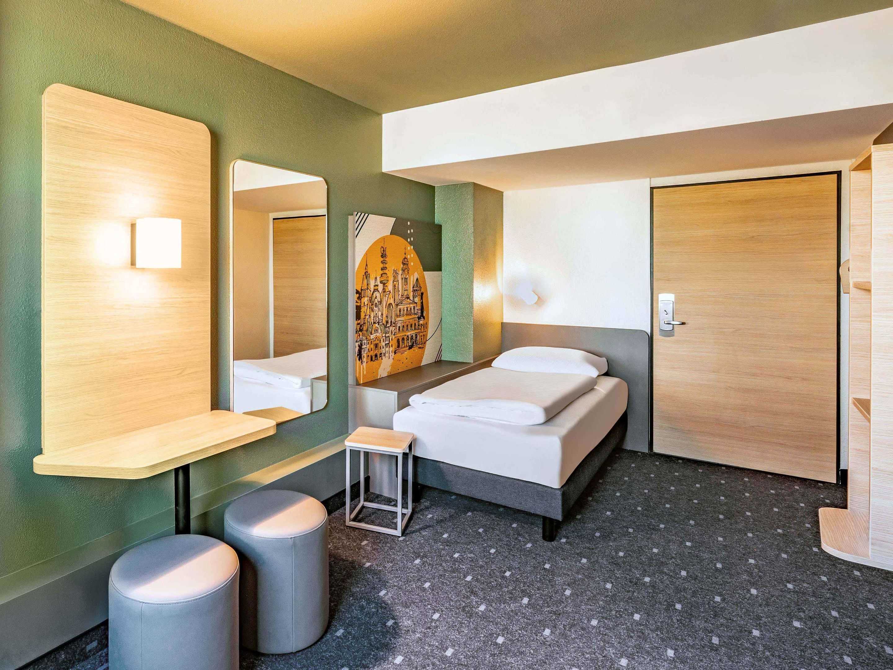 Una habitación de hotel moderna con una cama, un taburete pequeño, un espejo y un estante en la pared.