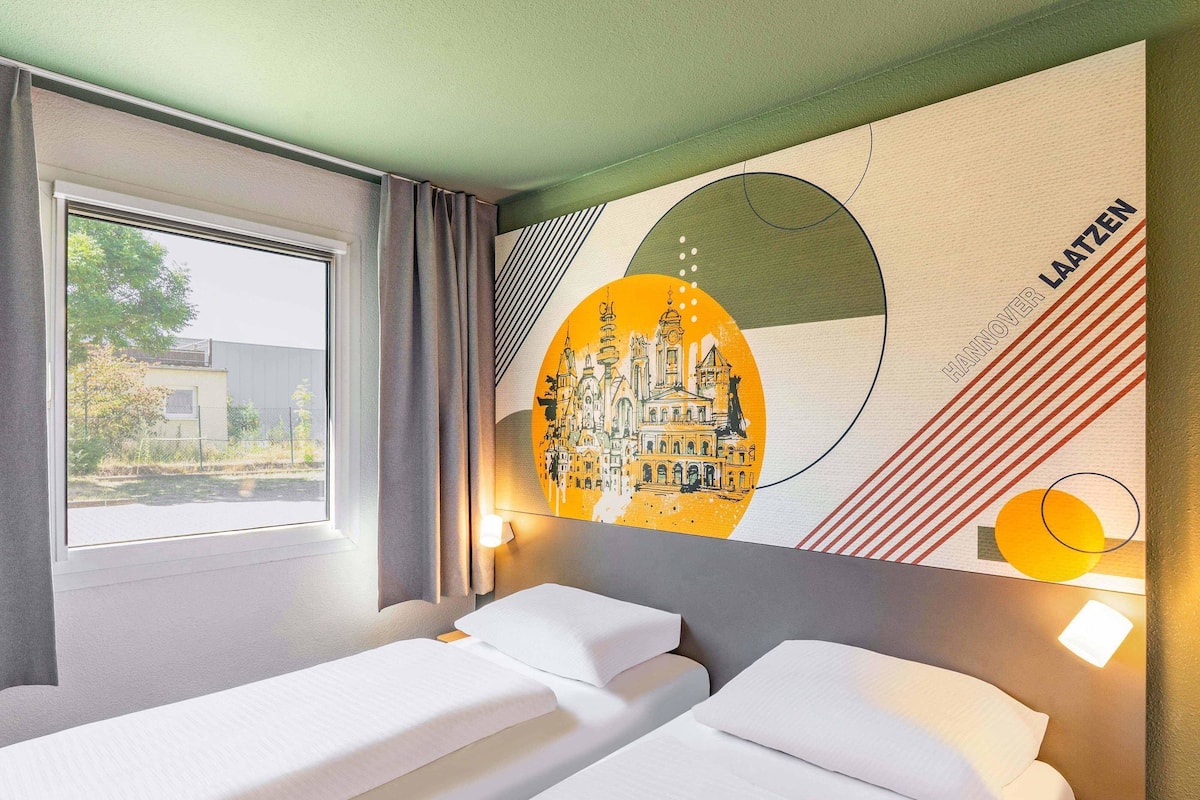 Een hotelkamer met twee bedden, een groot raam en een muurschildering van een stadsgezicht.