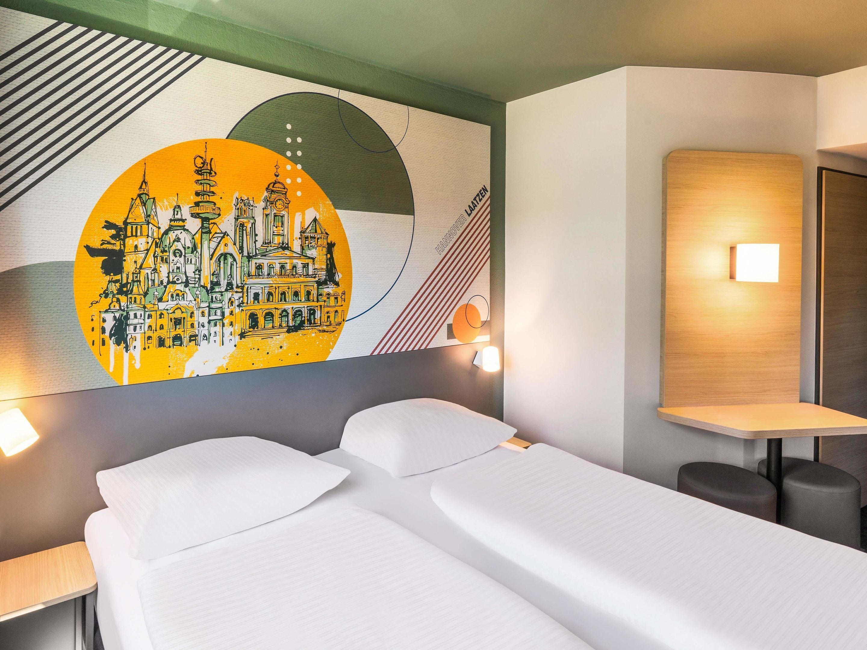 A hotel room with a bed, two bedside tables, a wall-mounted light, and a mural depicting a cityscape.