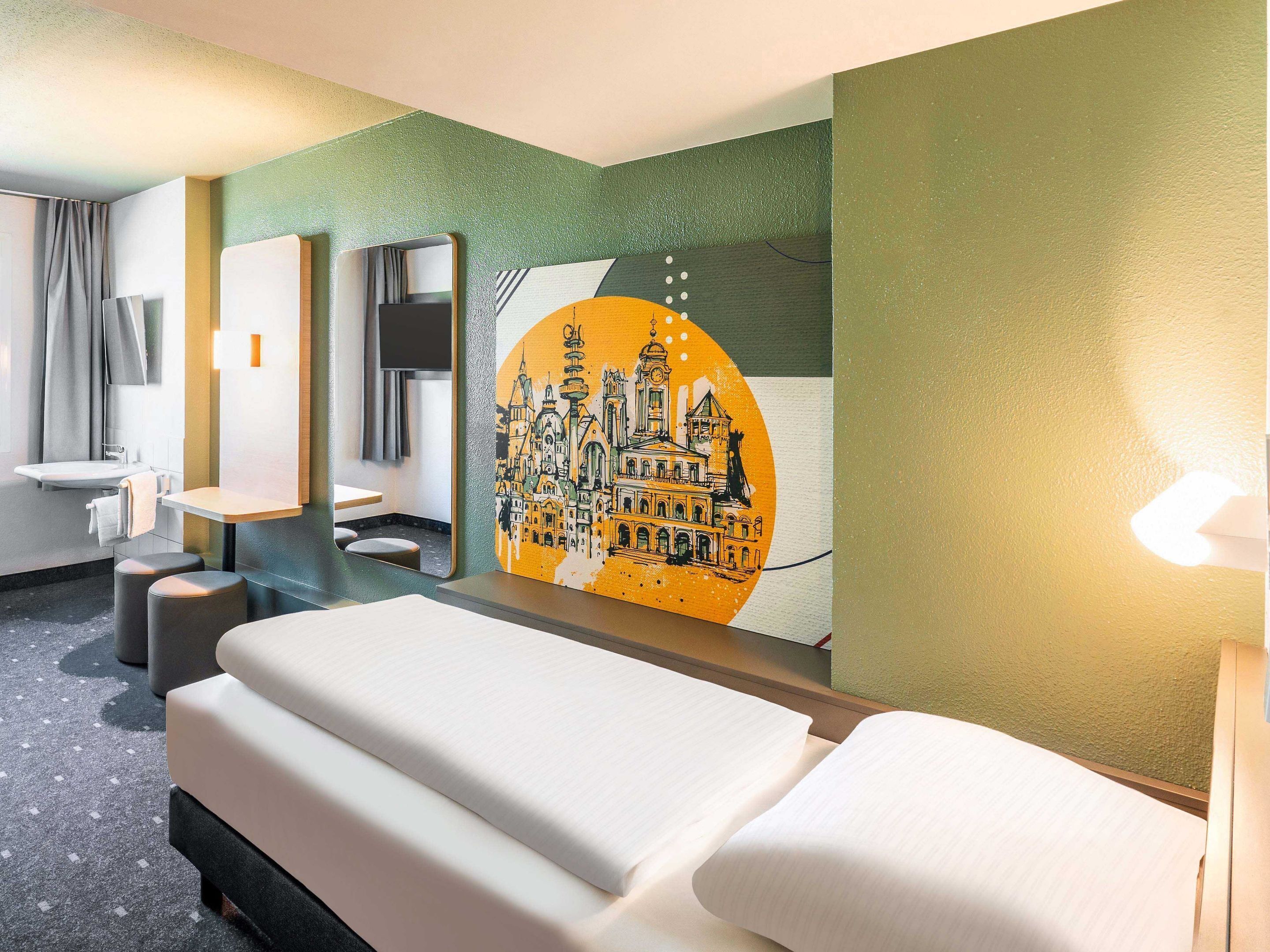 A hotel room with a bed, a desk, a chair, a mirror, and a wall with a cityscape painting.