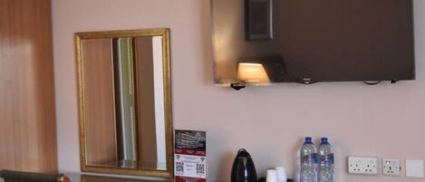 A hotel room with a large mirror, a flat-screen TV, a bedside table with a lamp, and a power outlet.