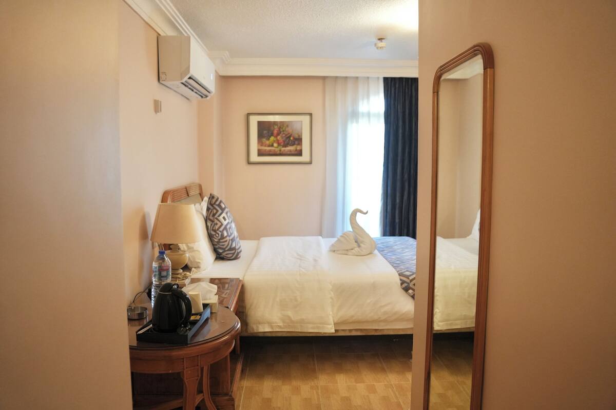 Ett hotellrum med en säng, ett skrivbord med en lampa, en vattenflaska, en kaffebryggare och en inramad tavla på väggen.