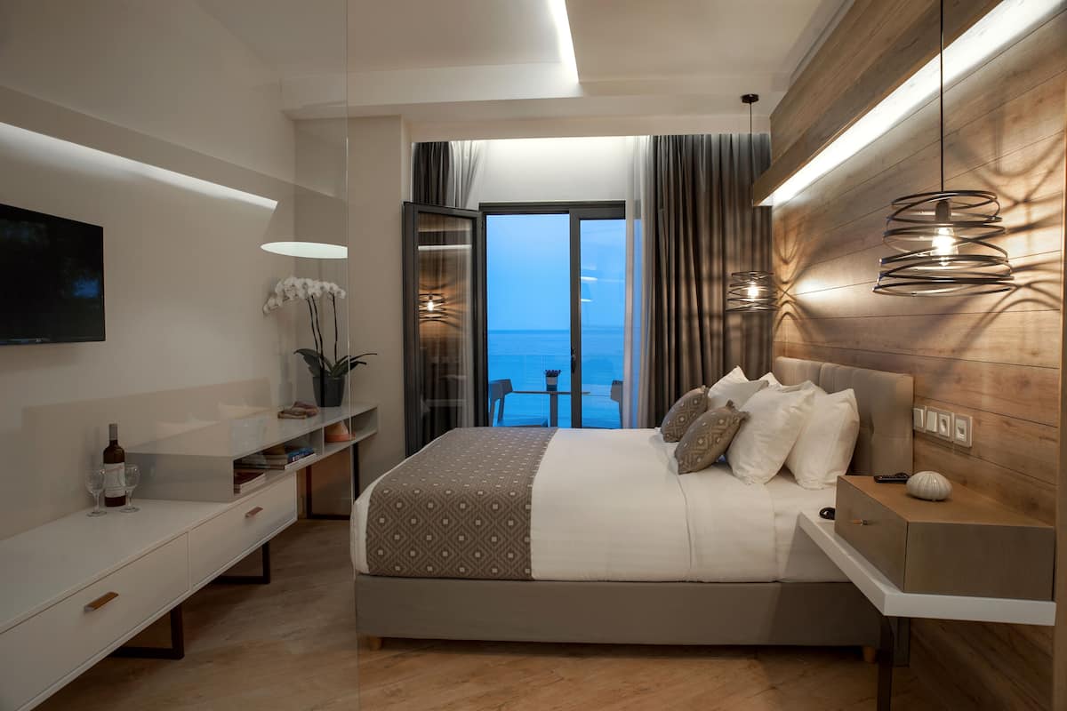 A modern bedroom with a large bed, a TV, a bedside table, and a view of the sea.