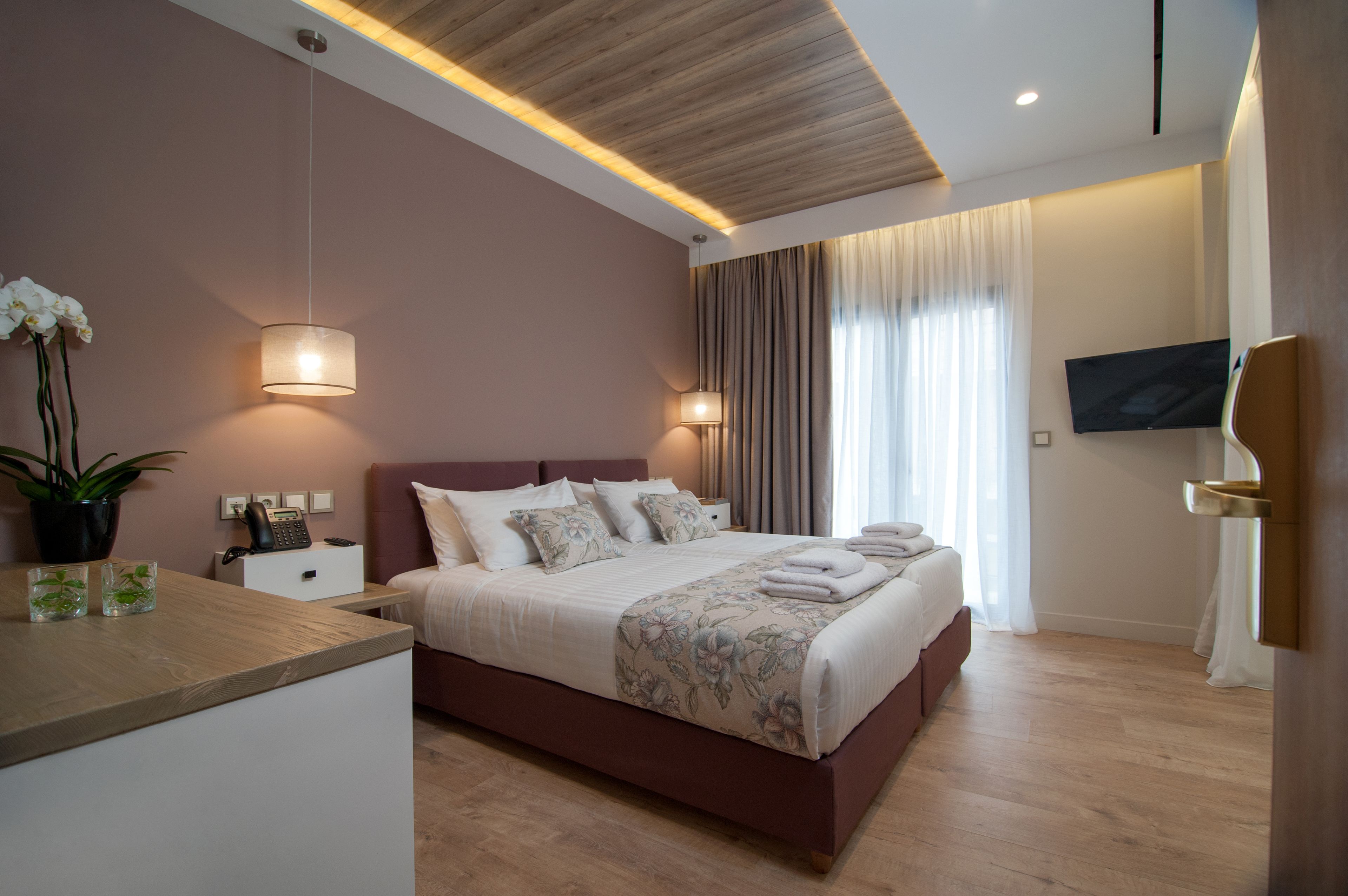 Un dormitorio moderno con techo de madera, una cama grande con ropa de cama floral, lámparas de noche, un televisor de pantalla plana y una planta en maceta.