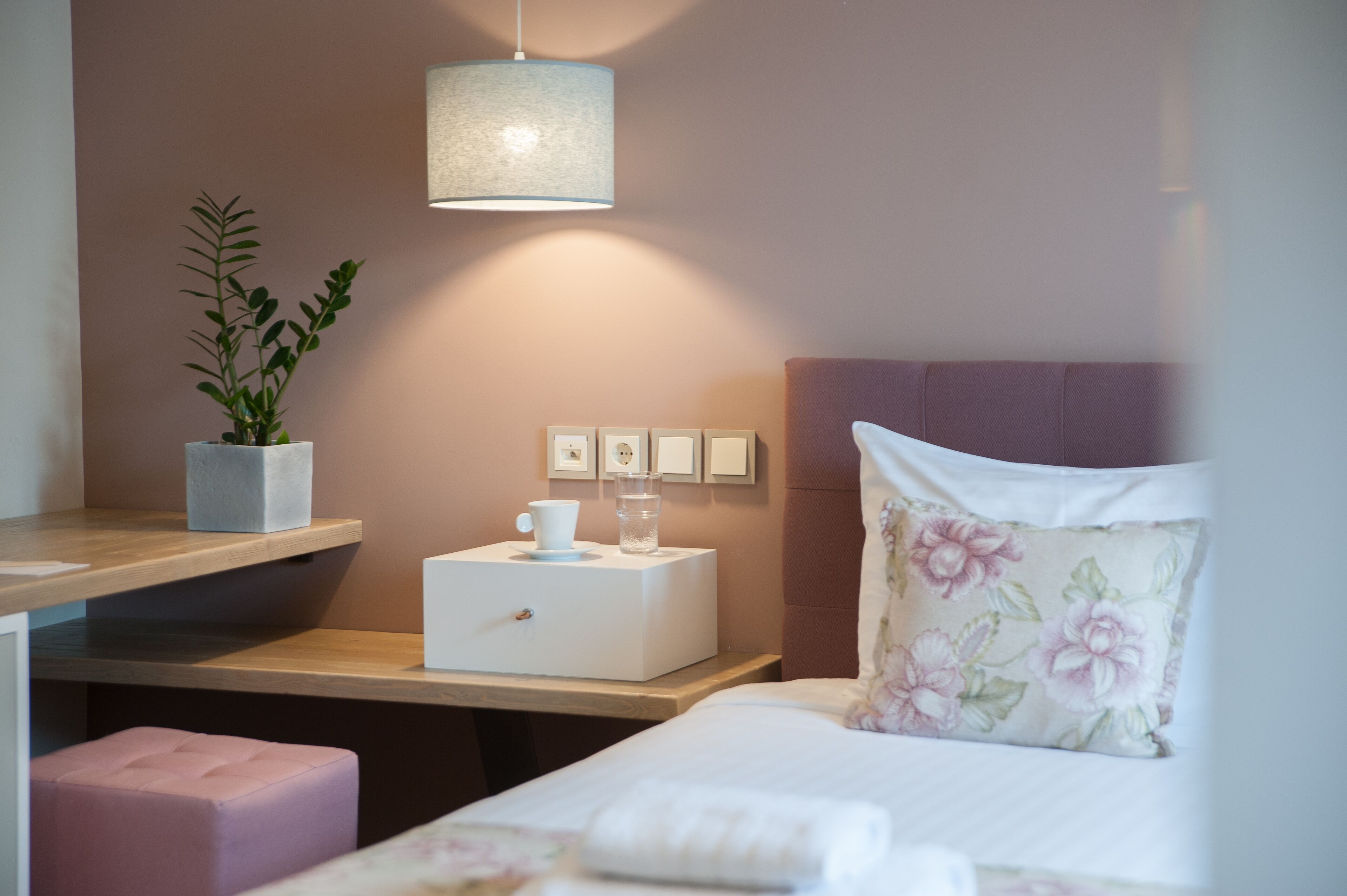 A hotel room with a bed, a bedside table with a lamp, a small plant, a water bottle, and a cup.