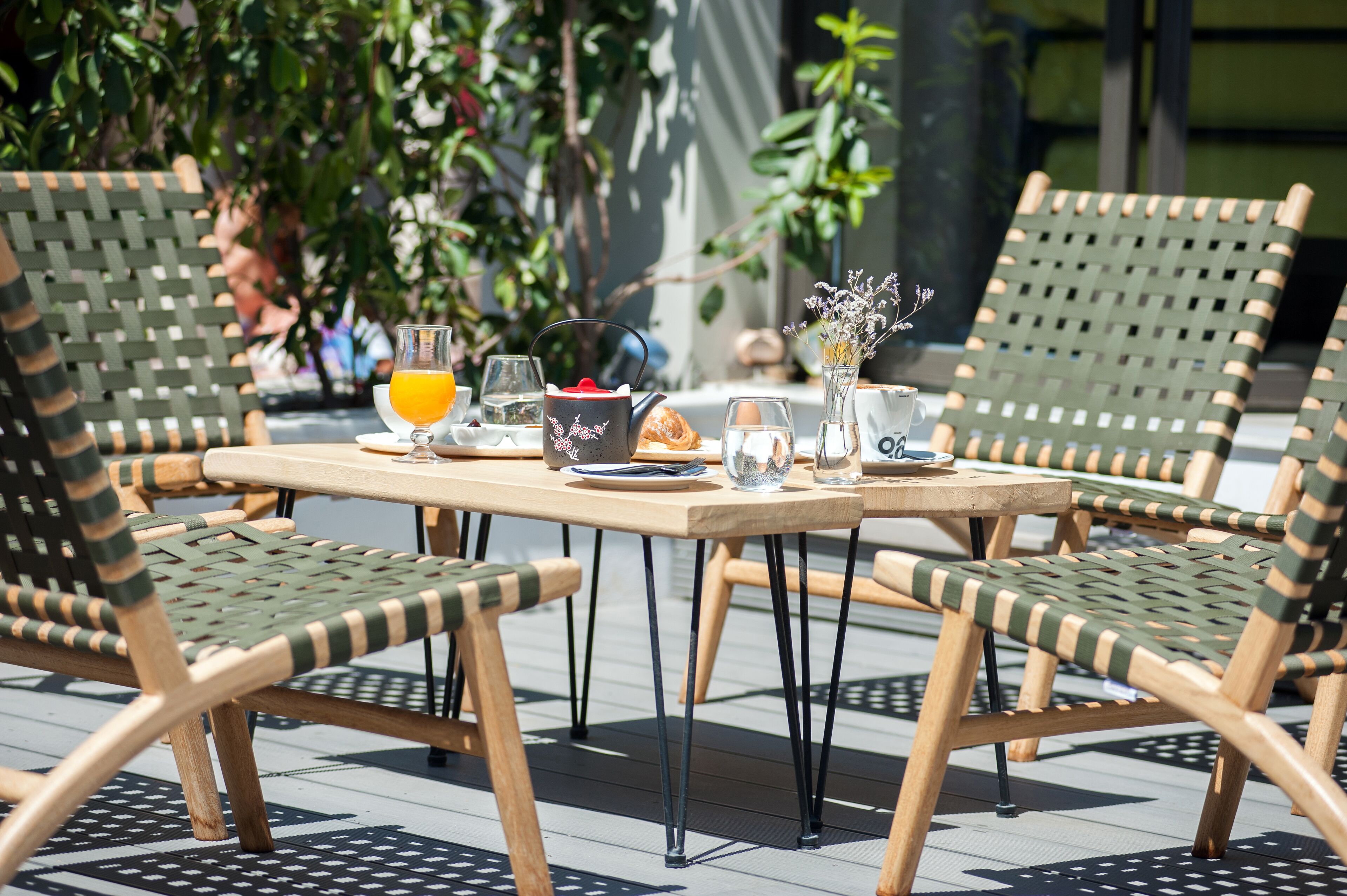 A table set outdoors with wicker chairs, a glass of orange juice, a bowl of fruit, and a vase with flowers.