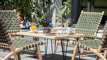 A table set outdoors with wicker chairs, a glass of orange juice, a bowl of fruit, and a vase with flowers.