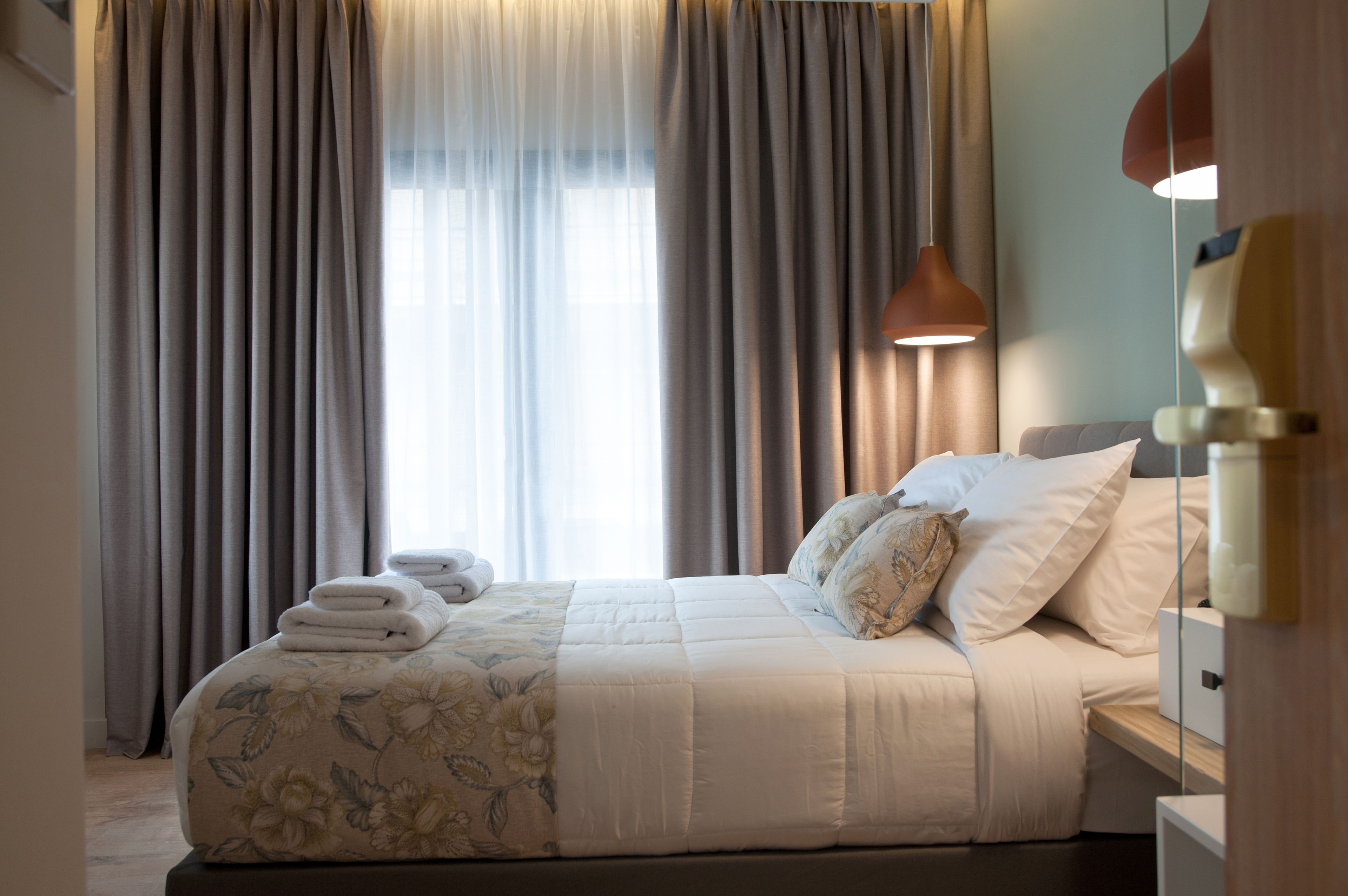 A neatly made bed with a floral duvet, a bedside lamp, and a stack of folded towels.