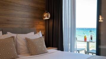 A hotel room with a wooden headboard, a bed with patterned pillows, a bedside table with a drink and a glass, and a view of the sea through large windows.
