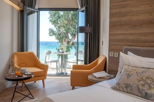 Habitación de hotel con una cama, dos sillones, una mesita con una bebida y un libro, y vista al mar a través de una puerta de cristal.