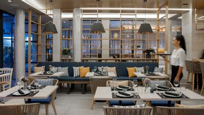 Un restaurante moderno con mobiliario de madera, cojines azules e iluminación colgante. Las mesas están dispuestas con platos, vasos y cubiertos.