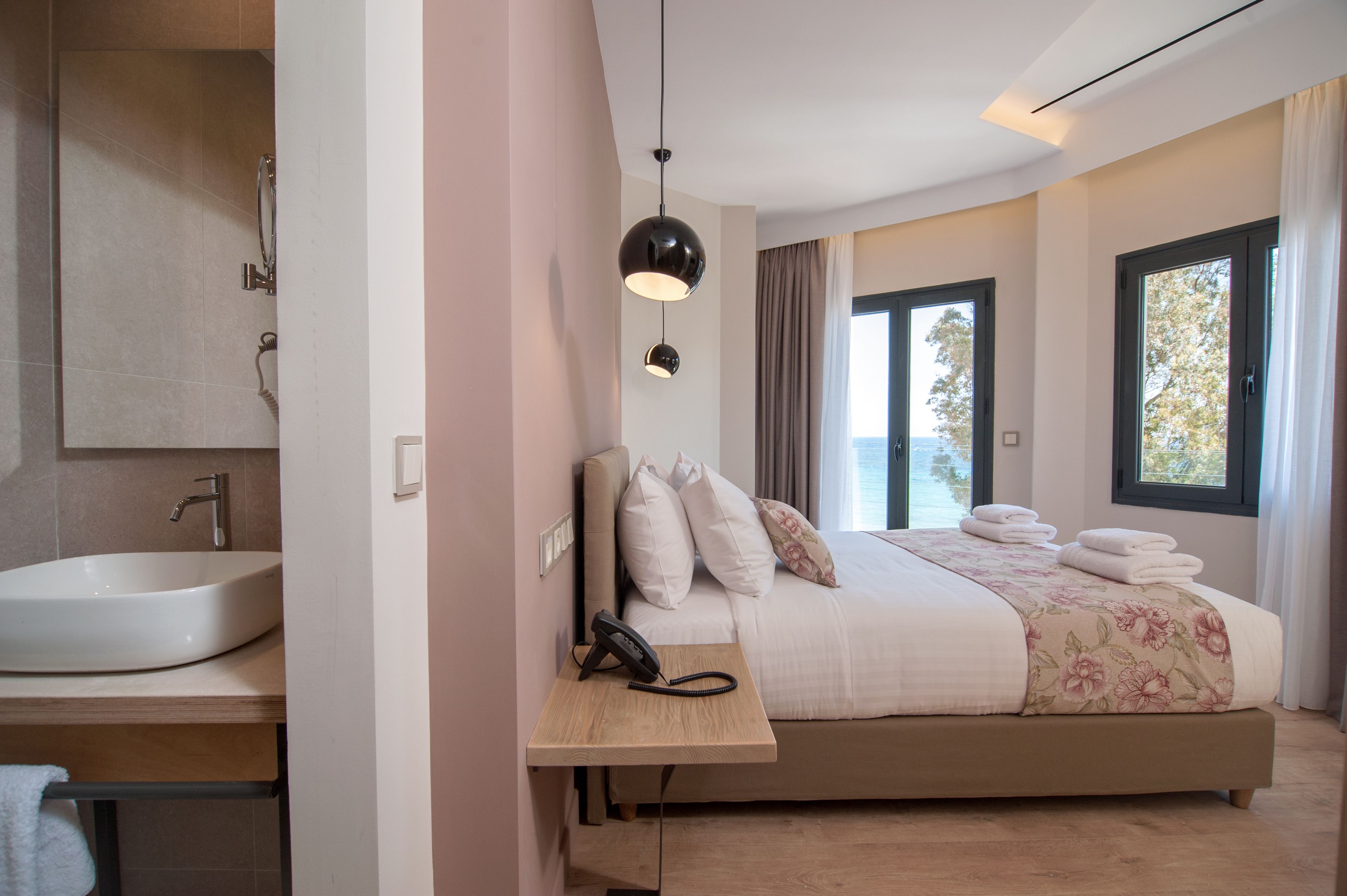 A modern hotel room with a large bed, a bedside table with a phone, a window with curtains, and a bathroom with a sink and shower.