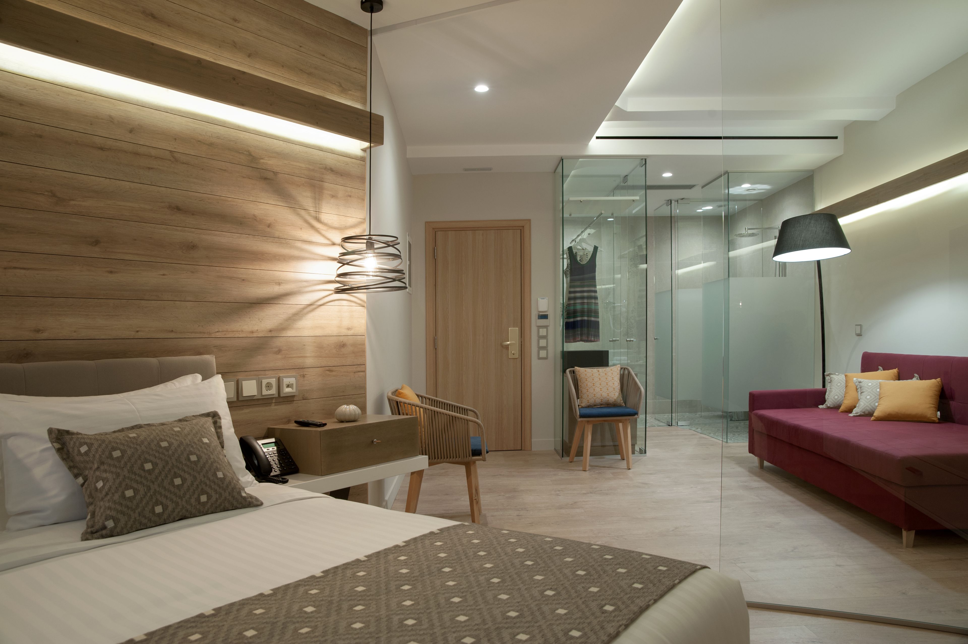 A modern bedroom with a wooden headboard, a bed with patterned pillows, a desk with a lamp, a red sofa, and a glass partition leading to a bathroom.