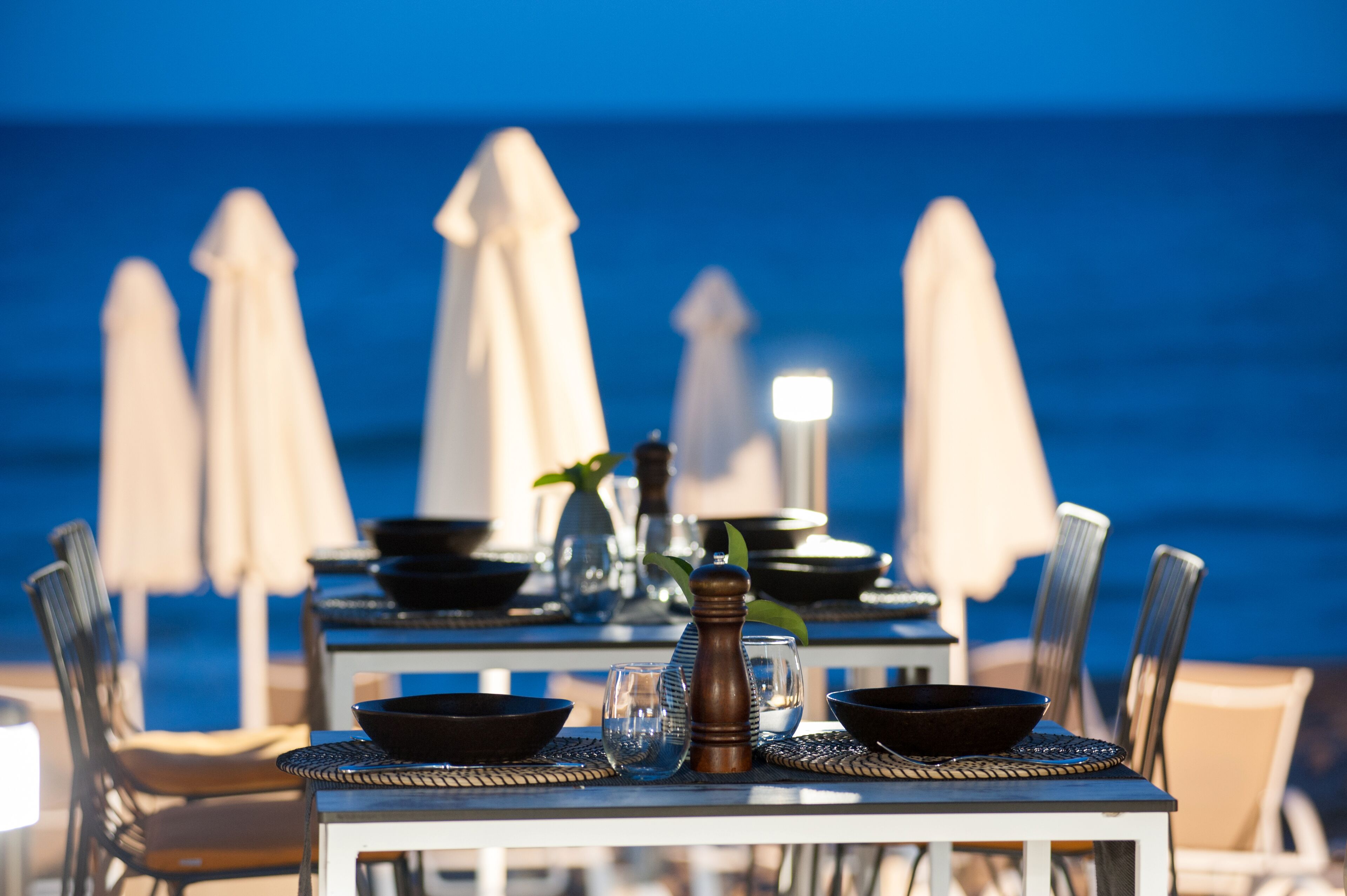 A table set for dining with a view of the ocean, white umbrellas, and chairs.
