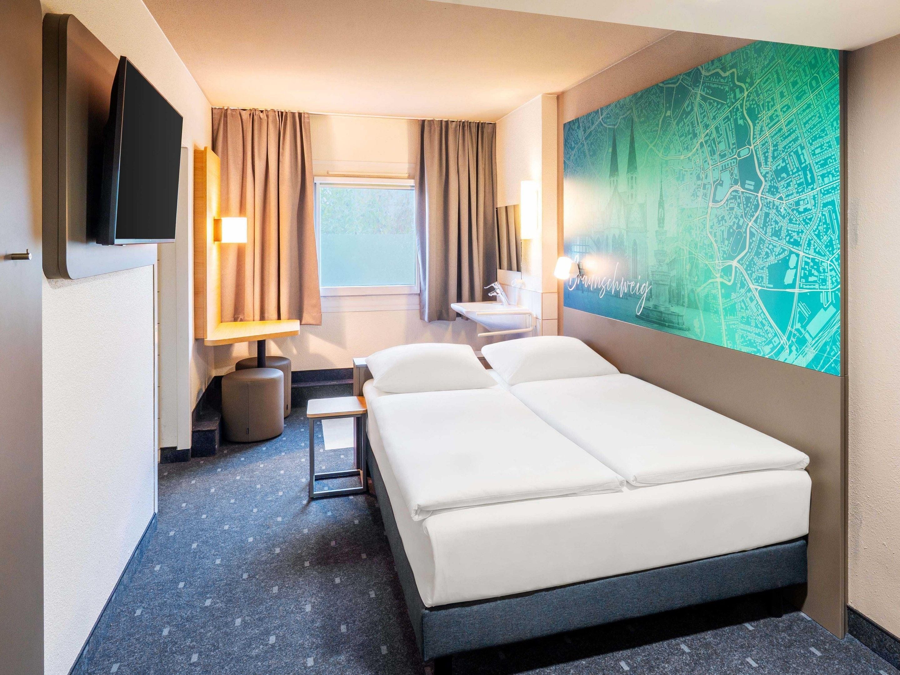 A hotel room with a bed, a desk, a chair, a lamp, a TV, a window with curtains, and a wall map.