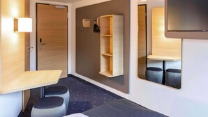 A modern hotel room with a wooden door, a small desk with a chair, a mirror, and a wall-mounted shelf. - B&B HOTEL Braunschweig-West (Braunschweig)