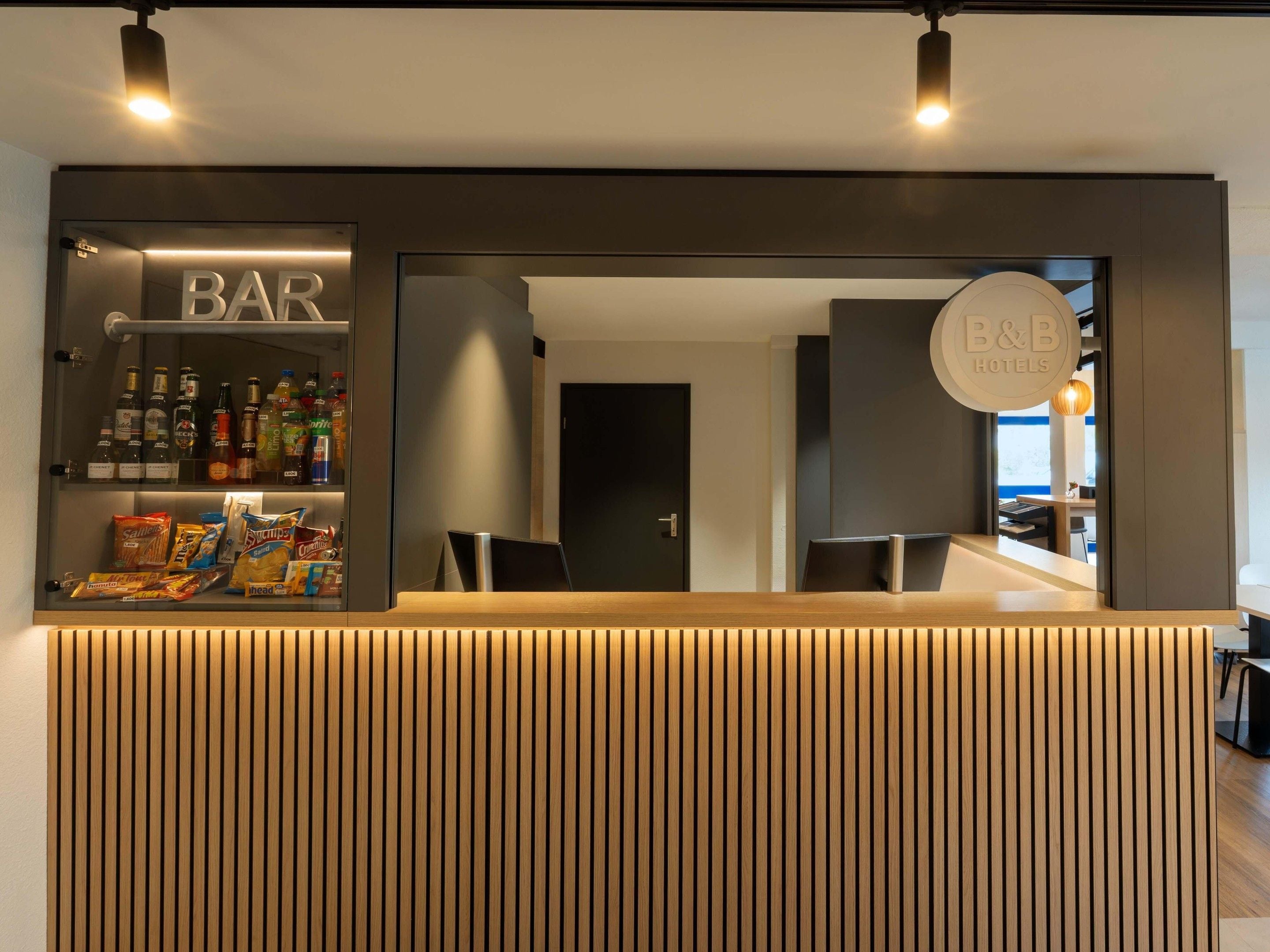 Een bar in een hotel, met een houten receptiebalie, planken vol snacks en drankjes, en een bord met de naam van het hotel.