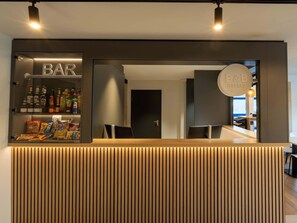 A hotel bar area with a wooden reception desk, shelves stocked with snacks and beverages, and a sign indicating the hotel's name.