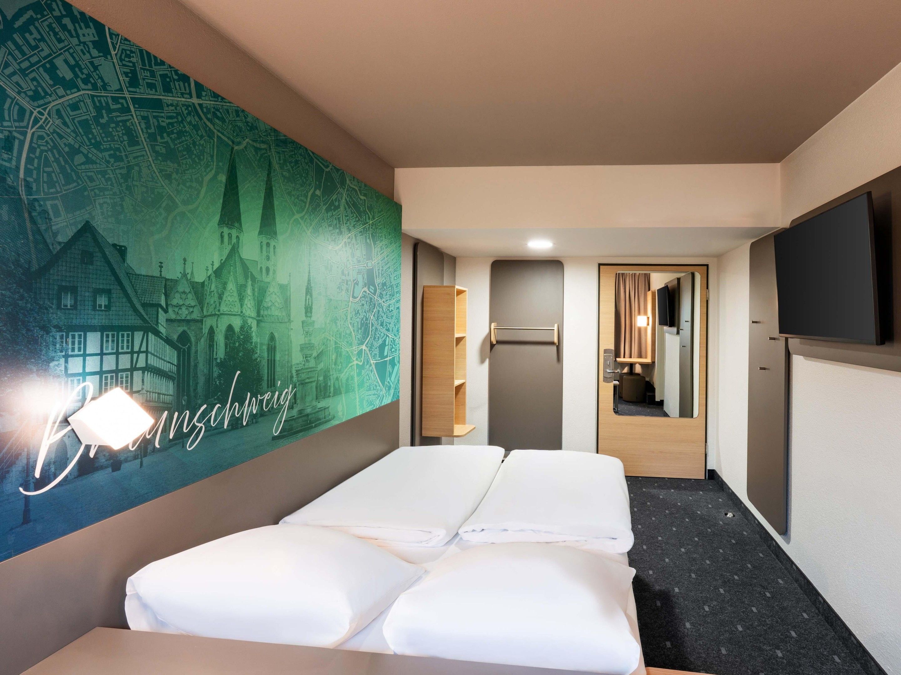 A hotel room with a bed, a flat-screen TV, a mirror, and a wall with a cityscape mural.